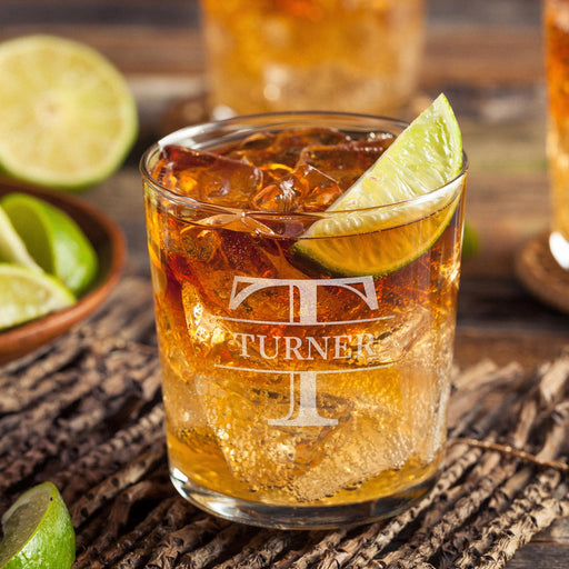 Personalized whiskey glass displayed with whiskey, ice, and fresh lime on a rustic wooden surface. The bold initial and name design is clearly visible, showing how the finished custom rocks glass looks in a home bar setting.