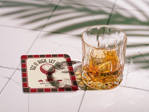 Custom square pulpboard coaster are shown laid out on marble floor. A whiskey glass is shown next to the coaster. The coaster features design with an illustration of a golf ball on a tee, checkerboard border, and custom text.
