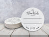 Front and back of coasters shown on a white wooden table against a blue wall. Front of coasters show the text I'm thankful for, has lines for writing in a response, and current year. Back of coasters feature a turkey illustration.