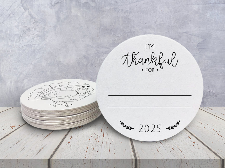 Front and back of coasters shown on a white wooden table against a blue wall. Front of coasters show the text I'm thankful for, has lines for writing in a response, and current year. Back of coasters feature a turkey illustration.