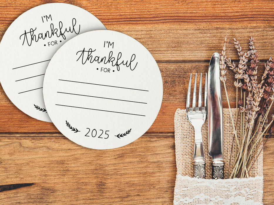 Front and back of coasters shown on an orange wooden surface next to a table setting. Front of coasters show the text I'm thankful for, has lines for writing in a response, and current year. Back of coasters feature a turkey illustration.