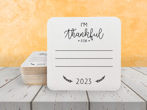 A stack of coasters by a single coaster on wooden table. Coasters shows the text I'm thankful for, has lines for writing in a response, and current year.