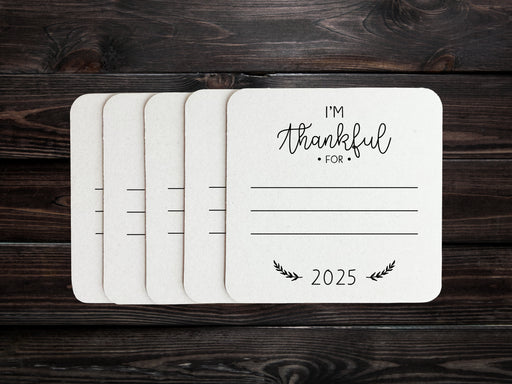 Square paper coasters are spread out on a dark wooden surface. Coasters feature the text I'm thankful for, has lines for writing in a response, and current year.