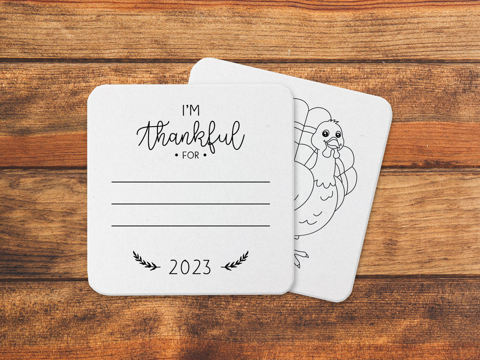 Front and back of coasters shown on an orange, wooden background. Front of coasters show the text I'm thankful for, has lines for writing in a response, and current year. Back of coasters feature a turkey illustration.