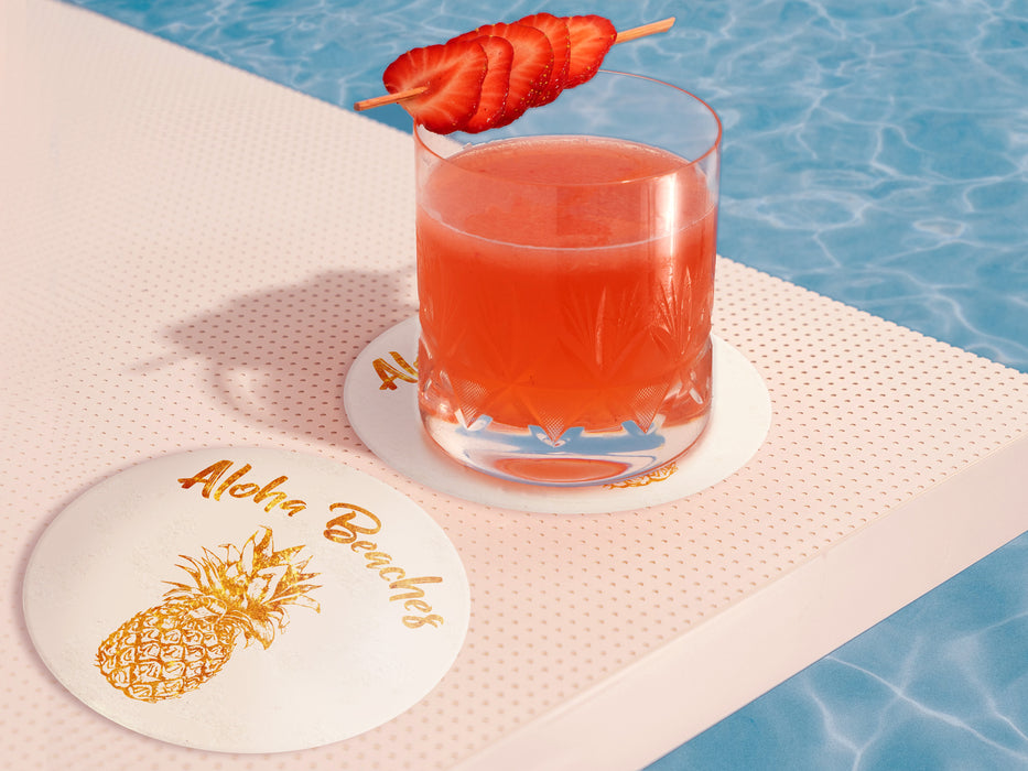 two coasters are shown on top of a stack of coasters. Coasters are white and feature Aloha Beaches design. Design is printed with a gold texture and shows the words Aloha Beaches and has a pineapple on it. Coasters are poolside with one under a short glass with a tropical pink red drink and strawberries