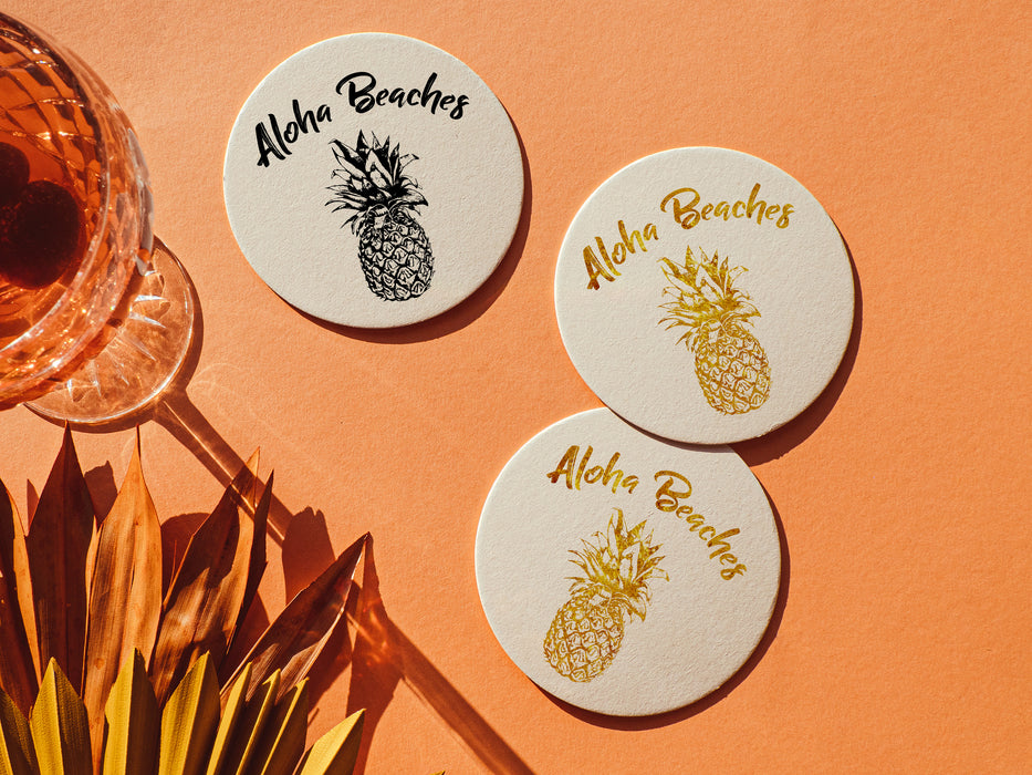 three white coasters are shown and feature Aloha Beaches design. Design is printed with a gold and black texture and shows the words Aloha Beaches and has a pineapple on it. Coasters are on orange background next to crystal glass with wine and tropical plants