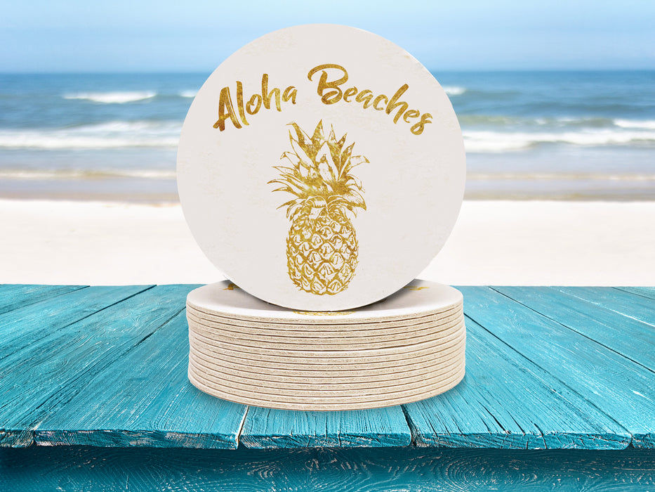 Single coaster is shown on top of a stack of coasters. Coasters are white and feature Aloha Beaches design. Design is printed with a gold texture and shows the words Aloha Beaches and has a pineapple on it ontop of blue wooden table with the ocean in the background