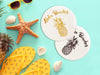 Two coasters are shown with a black and golf Aloha Beaches design. Design is printed with a gold and black texture and shows the words Aloha Beaches and has a pineapple on it. Coasters are on teal background with beach items around them such as shells, sea star, pineapple, sunglasses, and flip flops