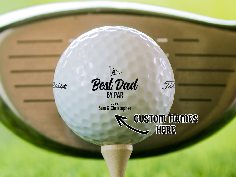 "Custom Names Here" single white titleist golf ball with customizable personalized black Best Dad By Par design on beige golf tee in front of golf club and golf course grass