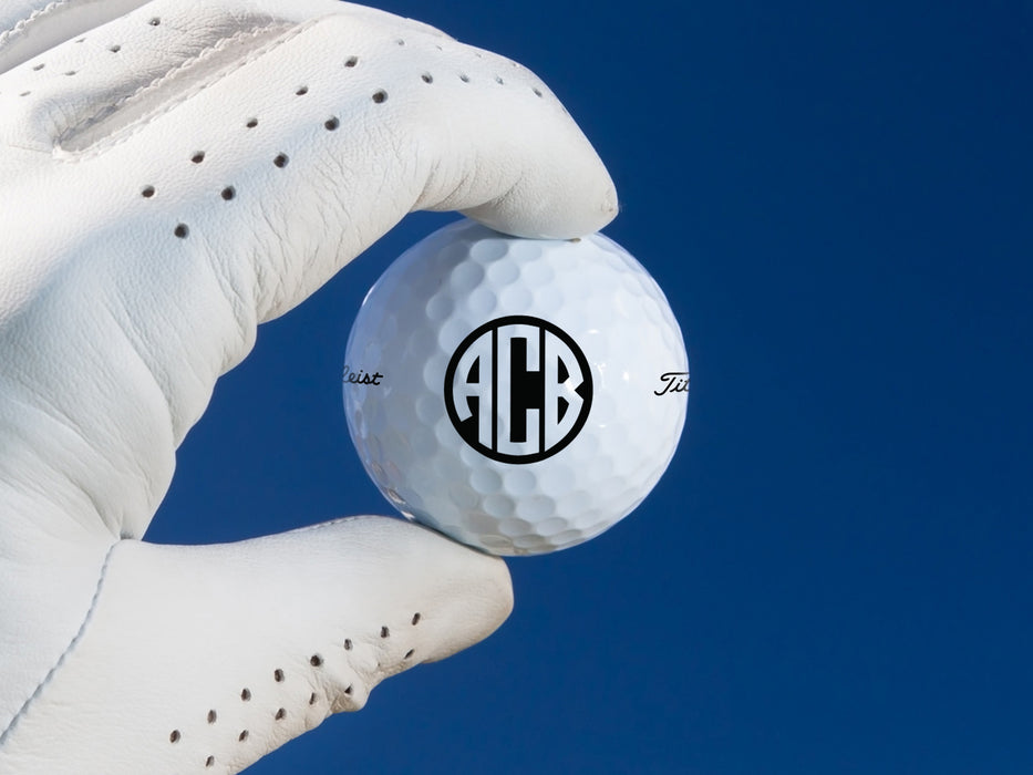 gloved hand holding single white titleist golf ball with custom personalized black monogram printed design against dark blue background