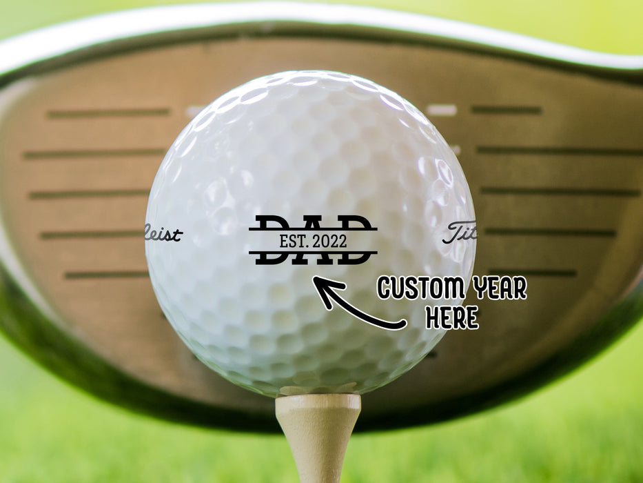 Custom Year Here: single white titleist golf ball with custom personalized established dad design on beige golf tee in front of golf club and golf course grass background