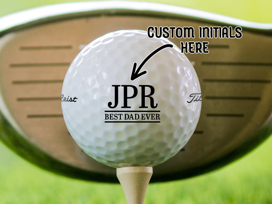 Custom Initials Here: single white titleist golf ball with customizable personalized initial Best Dad Ever design on beige golf tee in front of golf club and golf course grass