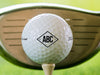 single white titleist golf ball with custom diamond monogram design on beige golf tee with golf course and golf club in the background