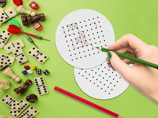 two white paper coasters with boxes and dots game design being drawn on with green and red colored pencils on green background. Coasters are surrounded by party toys such as chess pieces, dice, darts, etc.