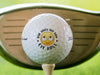 singe white titleist golf ball with dad jokes are how eye roll emoji design on beige golf tee in front of golf club and green grass course background