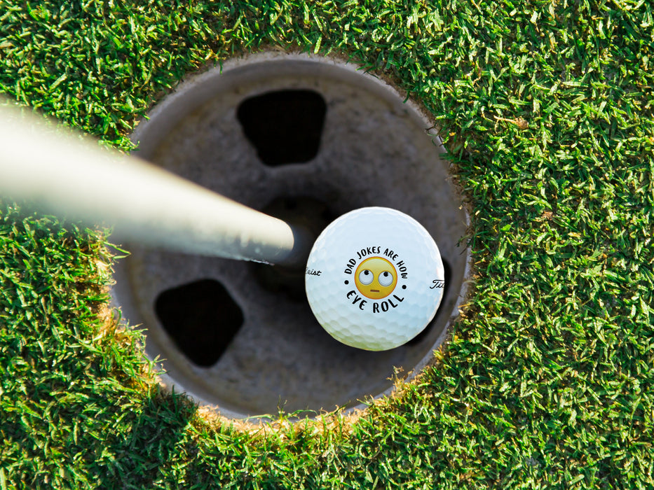 singe white titleist golf ball with dad jokes are how eye roll emoji design in golf course hole next to pole surrounded by green grass 