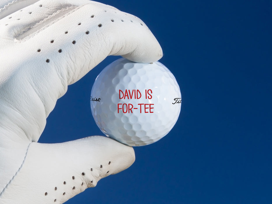 white gloved hand holding white titleist golf ball with type that says David is For-Tee on it against a dark blue background