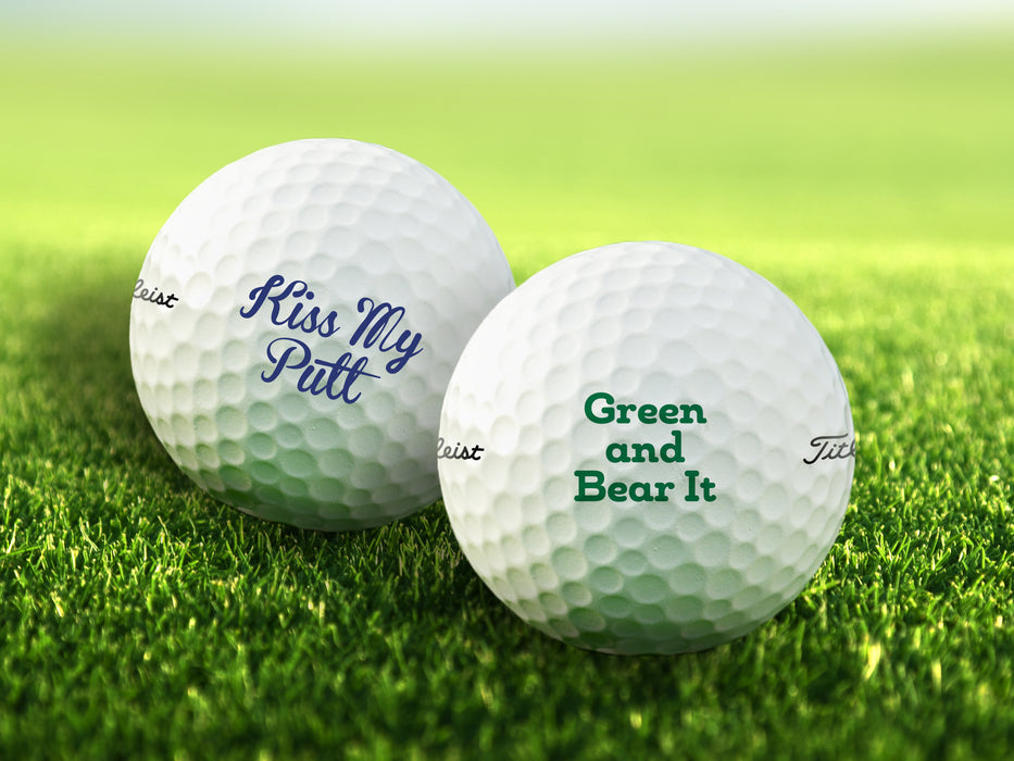 two white titleist golf balls with text that says Kiss my Putt and Green and Bear It on golf course grass