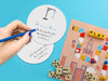 two white paper coasters with hangman design being drawn on by hand with blue colored pencils on bright blue background surrounded by toys and games such as dominos, board games, letter blocks, and game pieces