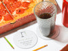 white table with pizza, soda can, and glass cup filled with soda next to white hangman party game coaster with writing on it next to two green and yellow colored pencils
