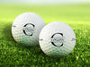 two white titleist golf balls with custom personalized black initial printed designs on grass golf course in the background