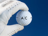 white gloved hand holding single white titleist golf ball with custom personalized black line monogram printed design against a blue background