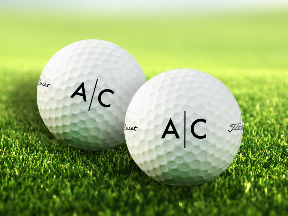 two white titleist golf balls with custom personalized black line monogram printed designs on golf course grass background