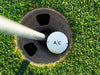 single white titleist golf ball with custom personalized black line monogram printed design in golf course hole next to pole surrounded by golf course grass