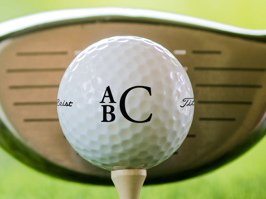 single white titleist golf ball with custom personalized black stacked monogram printed design in front of a golf club head with a grass golf course background