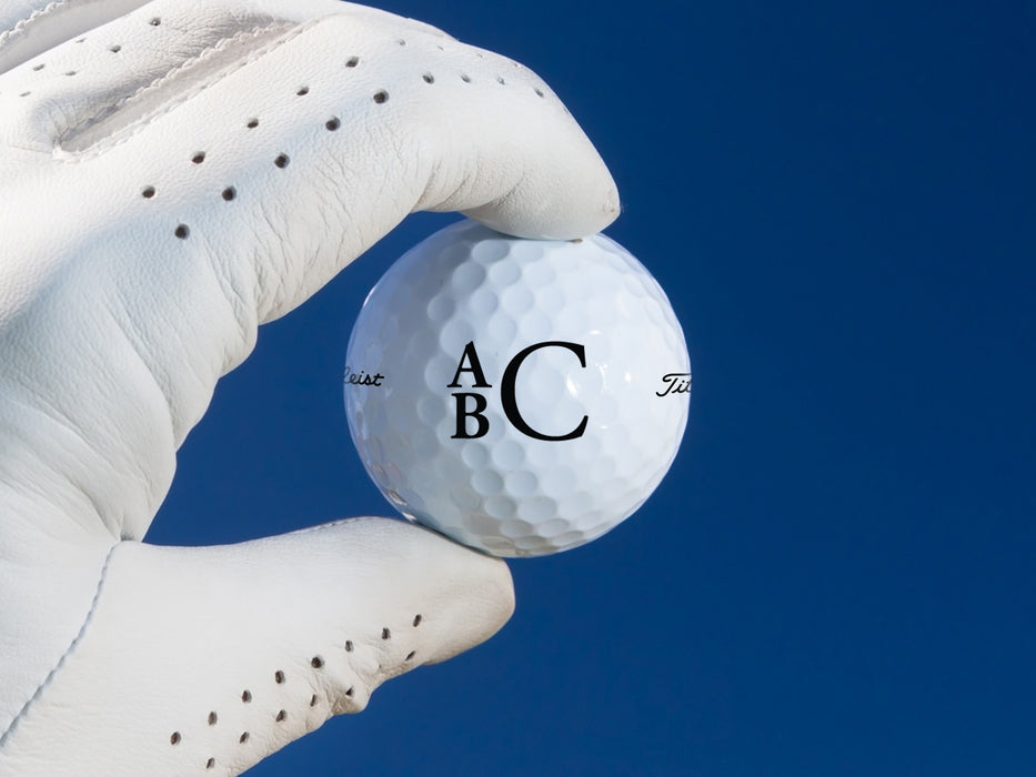 white gloved hand holding a single white titleist golf ball with custom personalized black stacked monogram printed design against a blue background