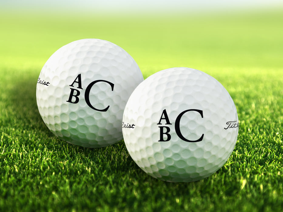 two white titleist golf balls with custom personalized black stacked monogram printed designs on a grass golf course background