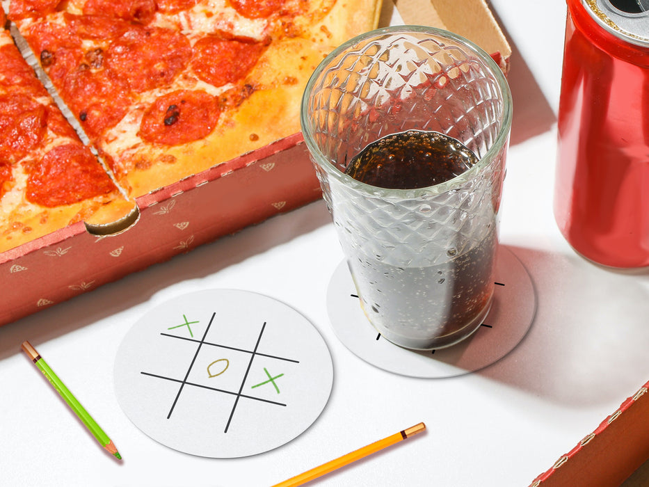 two white tic tac toe coasters on white table with pizza, soda can, and a glass cup filled with soda with green and yellow colored pencils