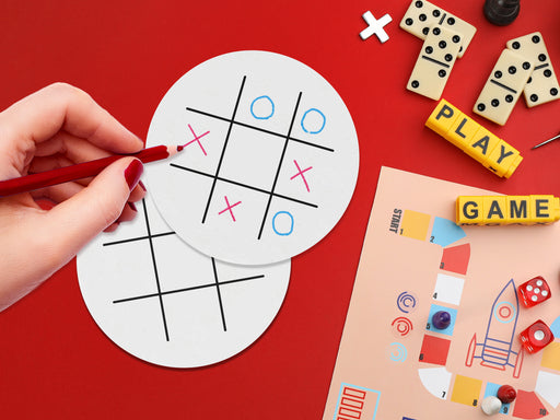 two white paper coasters with tic tac toe design on red background being drawn on with red colored pencil surrounded by game items such as letter blocks, board game, dominos, dice, and game pieces