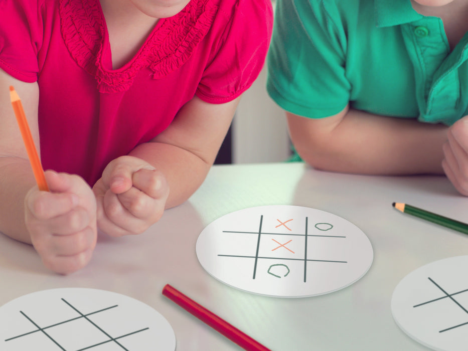 two kids in pink and green shirt playing with tic tac toe coaster game on white table with orange, green, and red colored pencils
