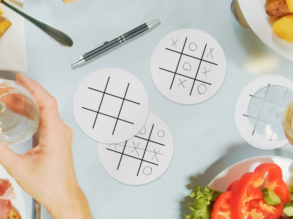 three white tic tac toe coasters with game drawn on them on blue dinner table surrounded by thanksgiving food and wine glasses as well as a black ballpoint pen