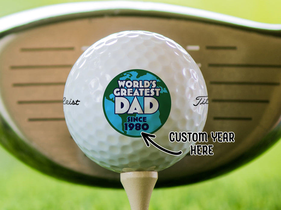 Custom Year Here: singe white titleist golf ball with customizable personalized World's Greatest Dad Globe Graphic with year design on beige golf tee in front of golf club and golf course grass in background