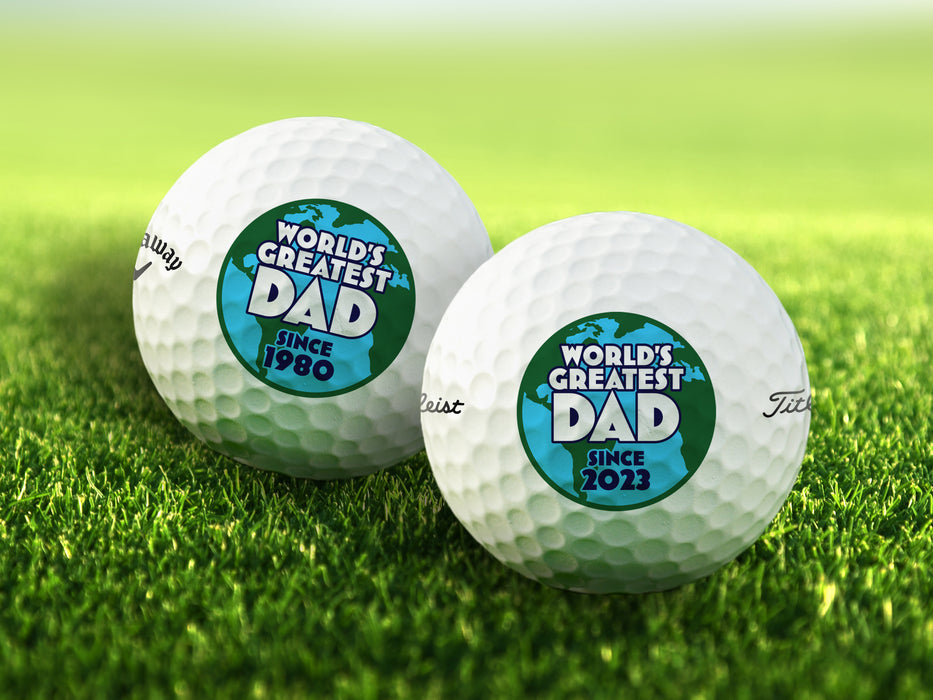 two white titleist golf balls with customizable personalized World's Greatest Dad Globe Graphic with year designs ontop of golf course grass