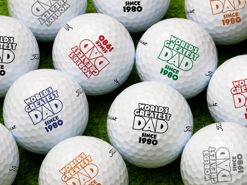Multiple white titleist golf balls with custom personalized World's Greatest Dad year designs in different colors on golf course grass