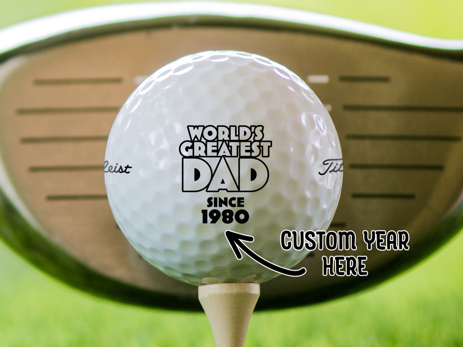 Custom Year Here single white titleist golf ball with custom personalized World's Greatest Dad year design on beige golf tee in front of golf club with grass background