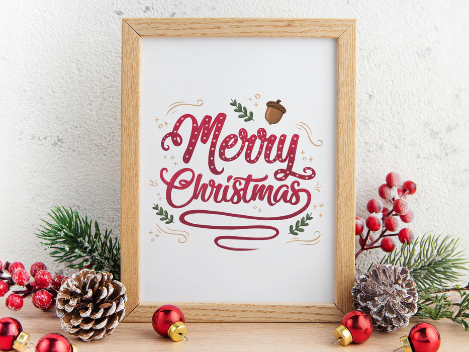 gold framed merry christmas typography print surrounded by holiday decor such as pinecones, pine leaves, ornaments, holly leaves