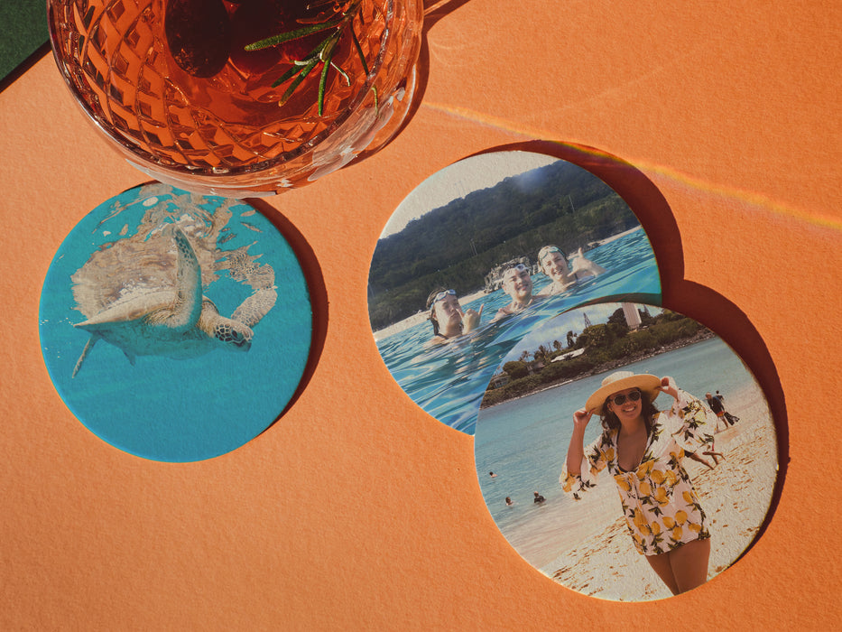 three paper photo coasters with pictures of people at the beach and a sea turtle on bright orange background under a glass of tropical drink with olives and rosemary in it