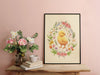 easter print of a baby chick surrounded by spring flowers in a black frame on a wooden table next to a vase of flowers and stacked books with a ceramic tea cup on top
