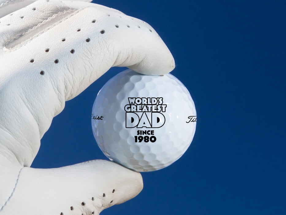 gloved hand holding single white titleist golf ball with custom personalized World's Greatest Dad year design  in front of dark blue background
