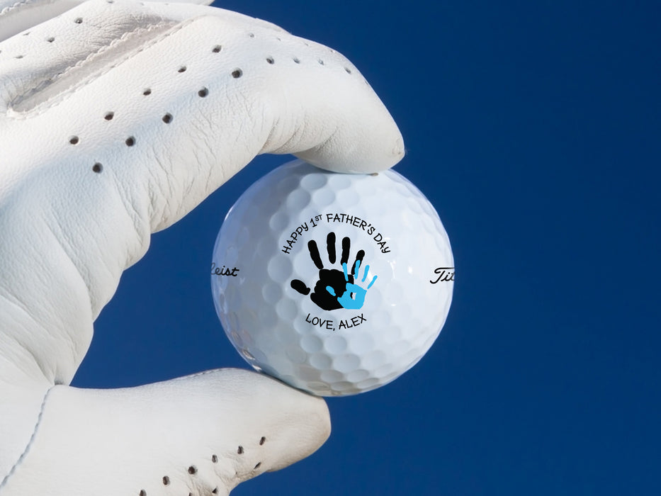 gloved hand holding single white titleist golf ball with custom personalized name happy 1st fathers day handprint design in front of dark blue background