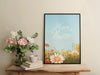happy easter print with a colorful meadow of spring flowers in a black frame sitting ontop of wooden table next to potted flowers and a book and ceramic tea cup