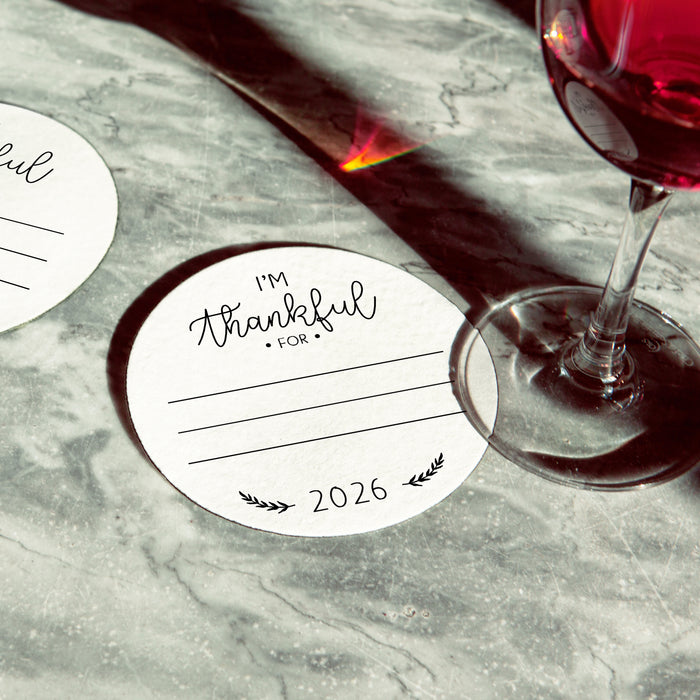 A white coaster says "I'm Thankful for" and "2026" on it, placed on a marble surface next to a glass of red wine.