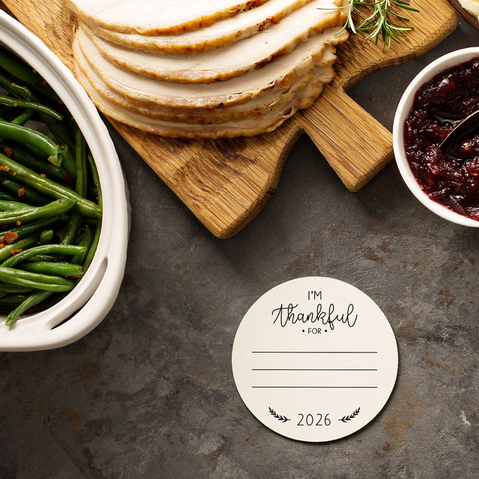 An off-white paper coaster with a Thanksgiving design on it sits on a Thanksgiving dinner table. The coasters features the words "I'm Thankful for" with lines beneath for someone to write what they are thankful for. The current year is also printed on the bottom.