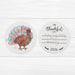 A round white coaster with a colorful illustration of a turkey and a handwritten message expressing gratitude.
