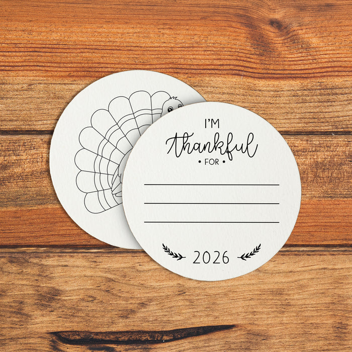 An off-white circular coaster with a turkey design and the text "I'm Thankful for" and "2026" printed on it. A wooden background is seen beneath the coasters.