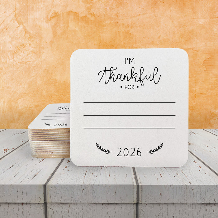 A white coaster with a message that says "I'm Thankful for" and "2026" printed on it, placed on a wooden surface. A stack of coasters with the same print are set behind the single coaster.